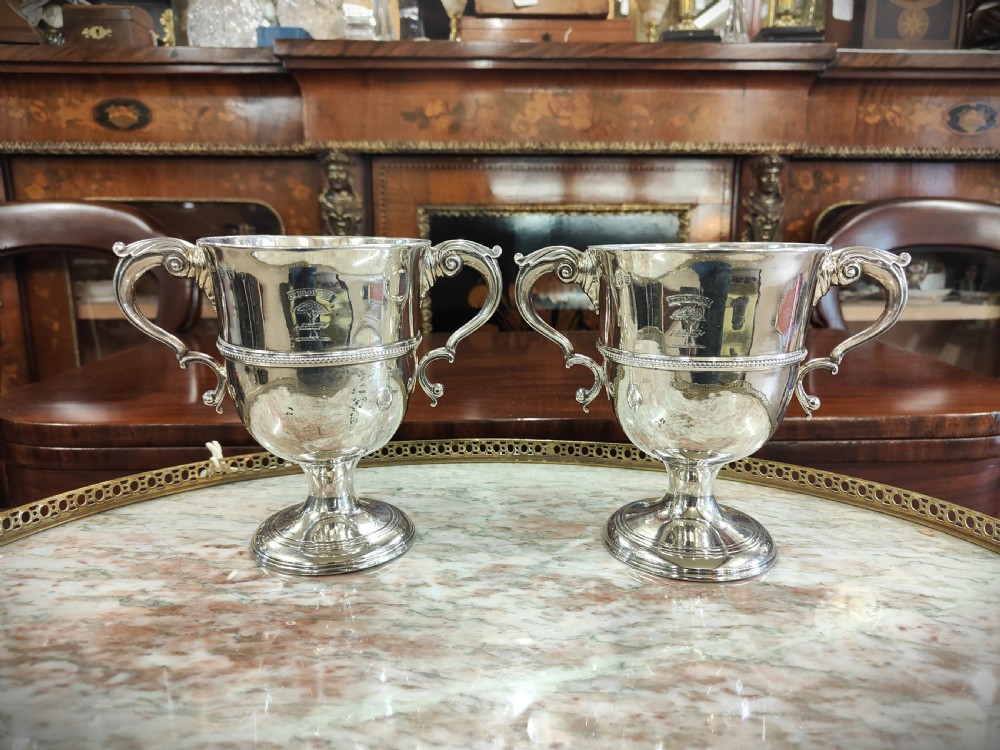 A pair of 18th-century Irish sterling silver twin-handled cups featuring central ribbed girdles and engraved crests on pedestal bases.