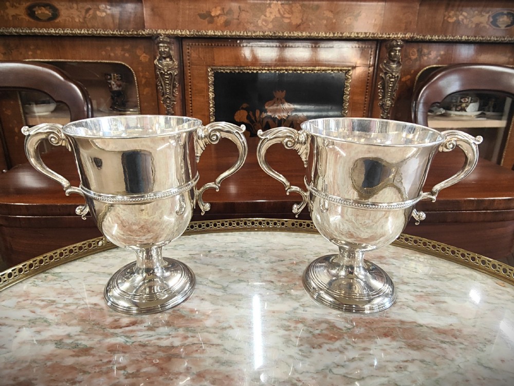 A pair of 18th-century Irish sterling silver twin-handled cups featuring central ribbed girdles and engraved crests on pedestal bases. View II