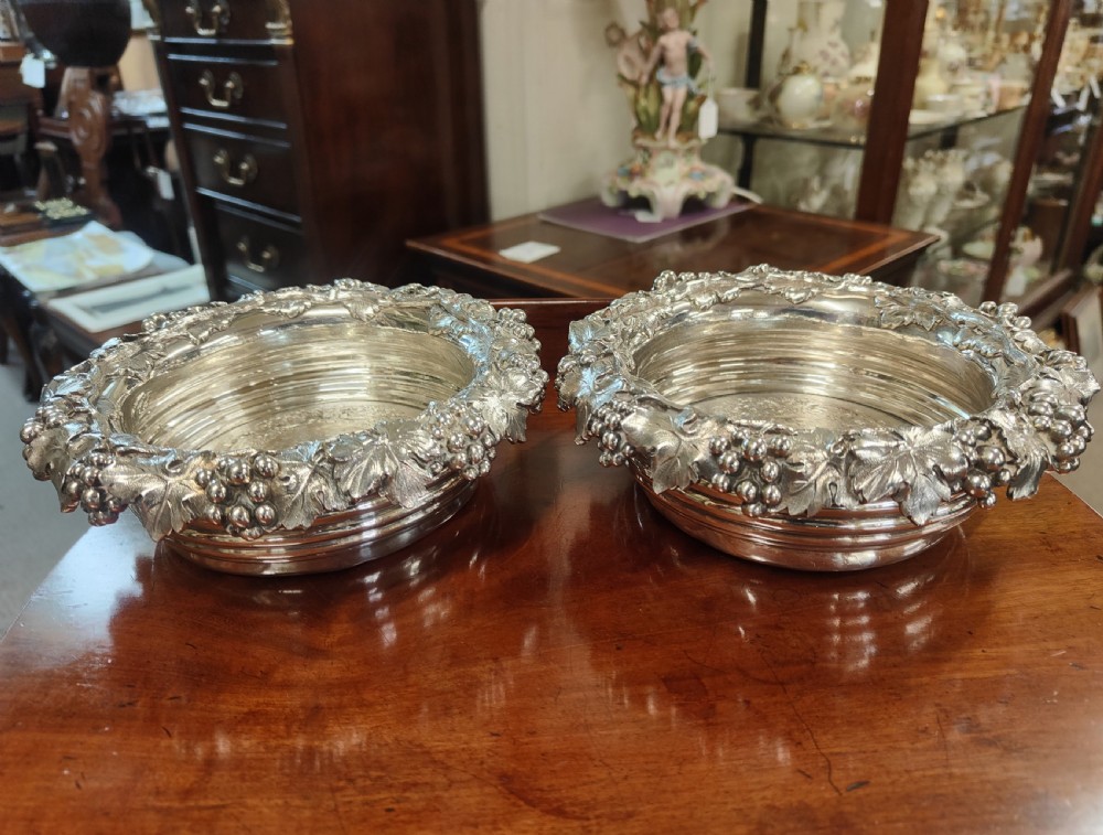 A pair of circular Georgian silver wine coasters featuring intricate cast grape and vine leaf borders with turned wooden bases.