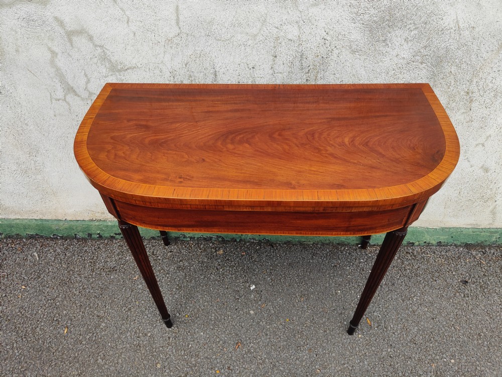 A Georgian mahogany demilune card table featuring satinwood crossbanding along the folded top and fluted tapered legs.