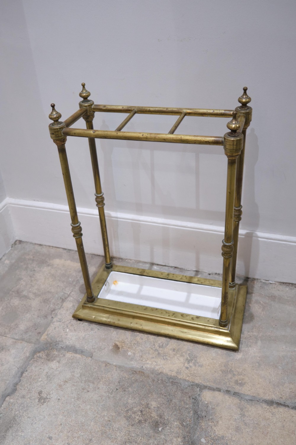 A Victorian brass umbrella stand featuring turned uprights with finials, a divided top rail, and a rectangular white drip tray base.