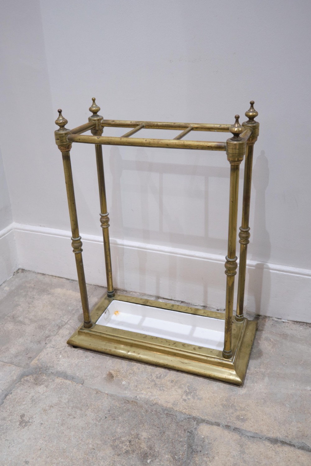 A Victorian brass umbrella stand featuring turned uprights with finials, a divided top rail, and a rectangular white drip tray base. View II