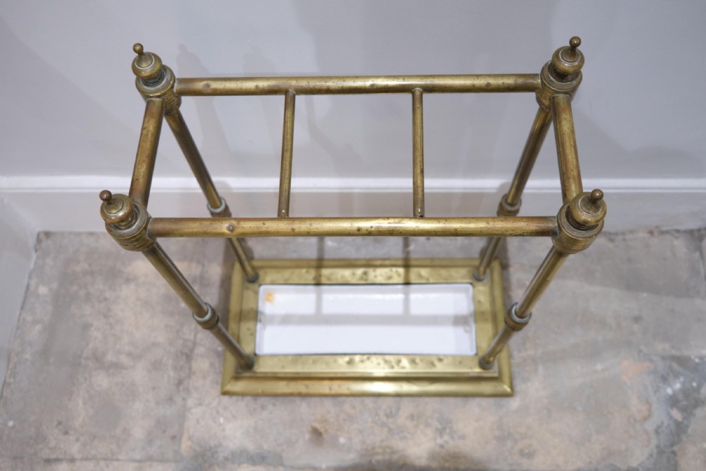 A Victorian brass umbrella stand featuring turned uprights with finials, a divided top rail, and a rectangular white drip tray base. View V