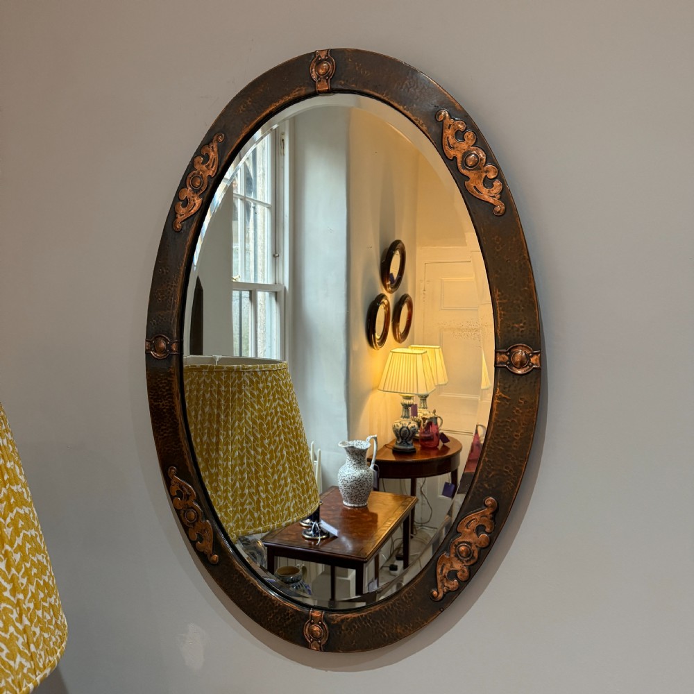 An oval Arts and Crafts wall mirror featuring a hand-hammered copper frame decorated with stylised scrollwork and circular bosses.