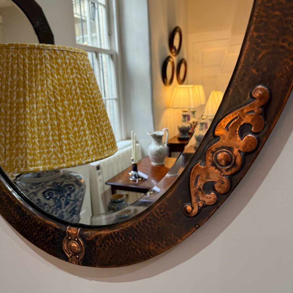 An oval Arts and Crafts wall mirror featuring a hand-hammered copper frame decorated with stylised scrollwork and circular bosses. View III