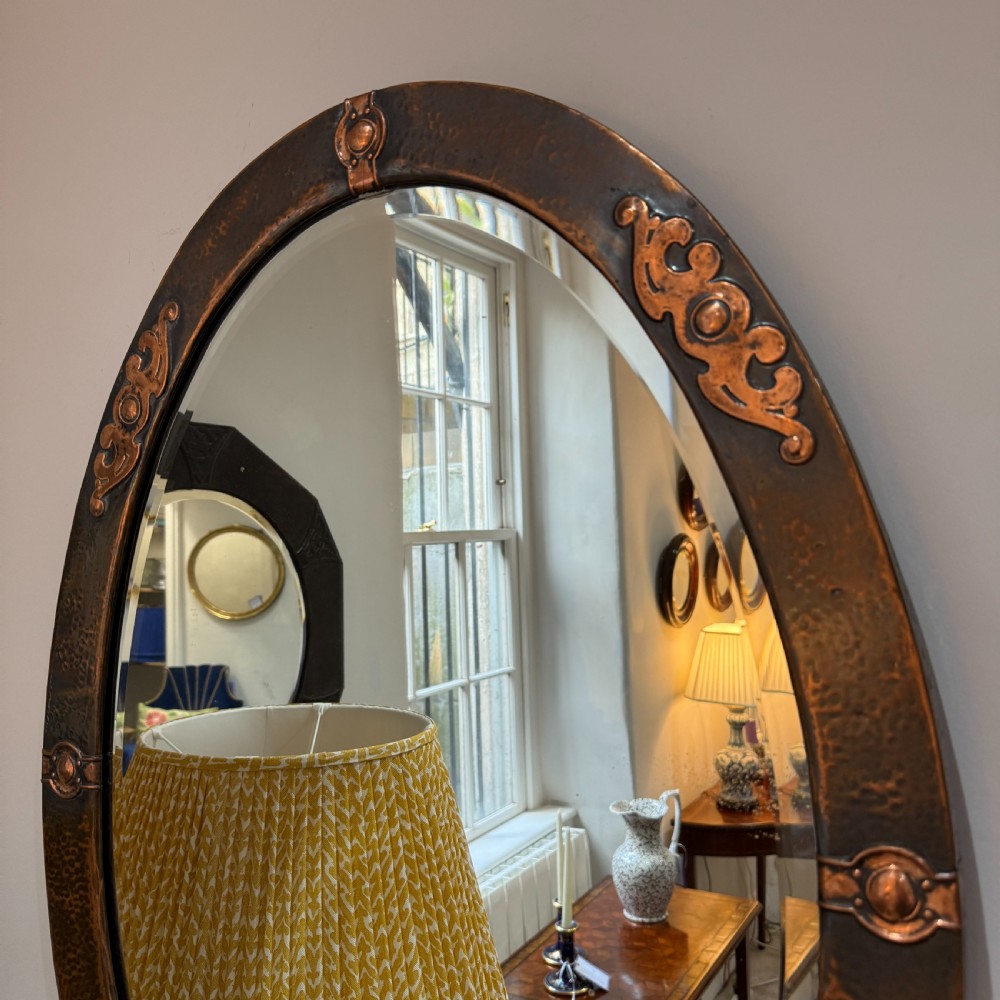 An oval Arts and Crafts wall mirror featuring a hand-hammered copper frame decorated with stylised scrollwork and circular bosses. View IV