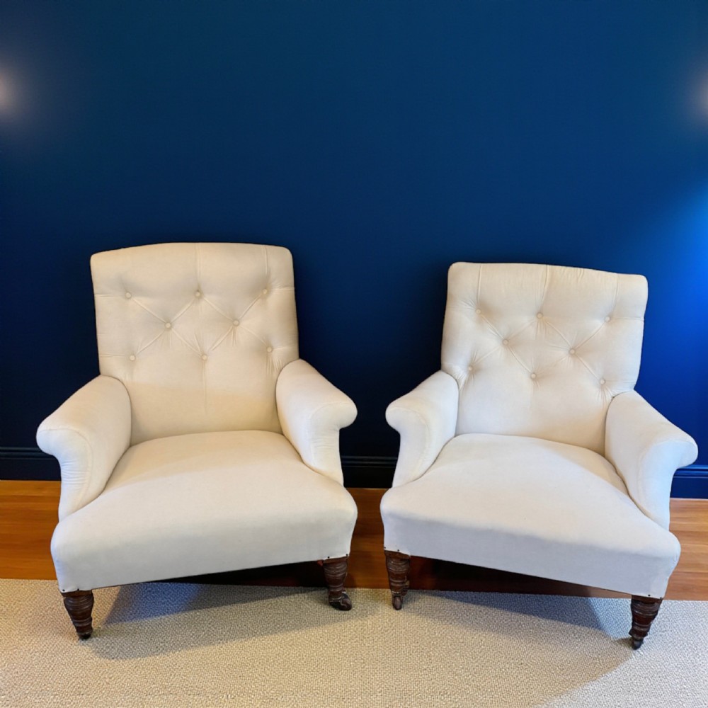 A pair of 19th-century Howard-style armchairs upholstered in cream fabric featuring deep button-backed tufting and turned dark walnut front legs on brass castors.