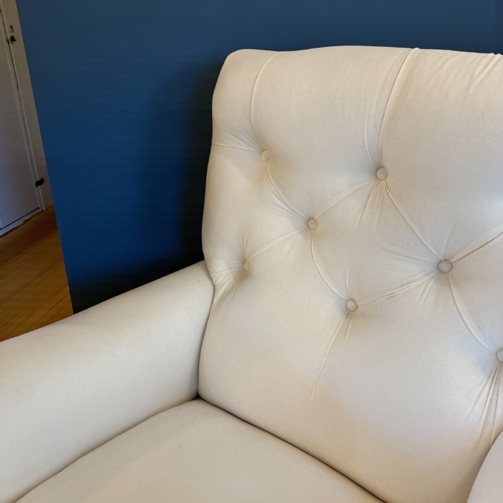 A pair of 19th-century Howard-style armchairs upholstered in cream fabric featuring deep button-backed tufting and turned dark walnut front legs on brass castors. View III