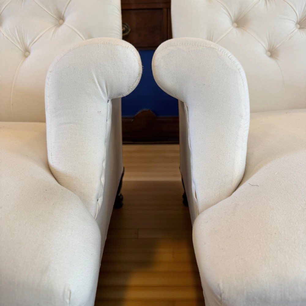 A pair of 19th-century Howard-style armchairs upholstered in cream fabric featuring deep button-backed tufting and turned dark walnut front legs on brass castors. View V