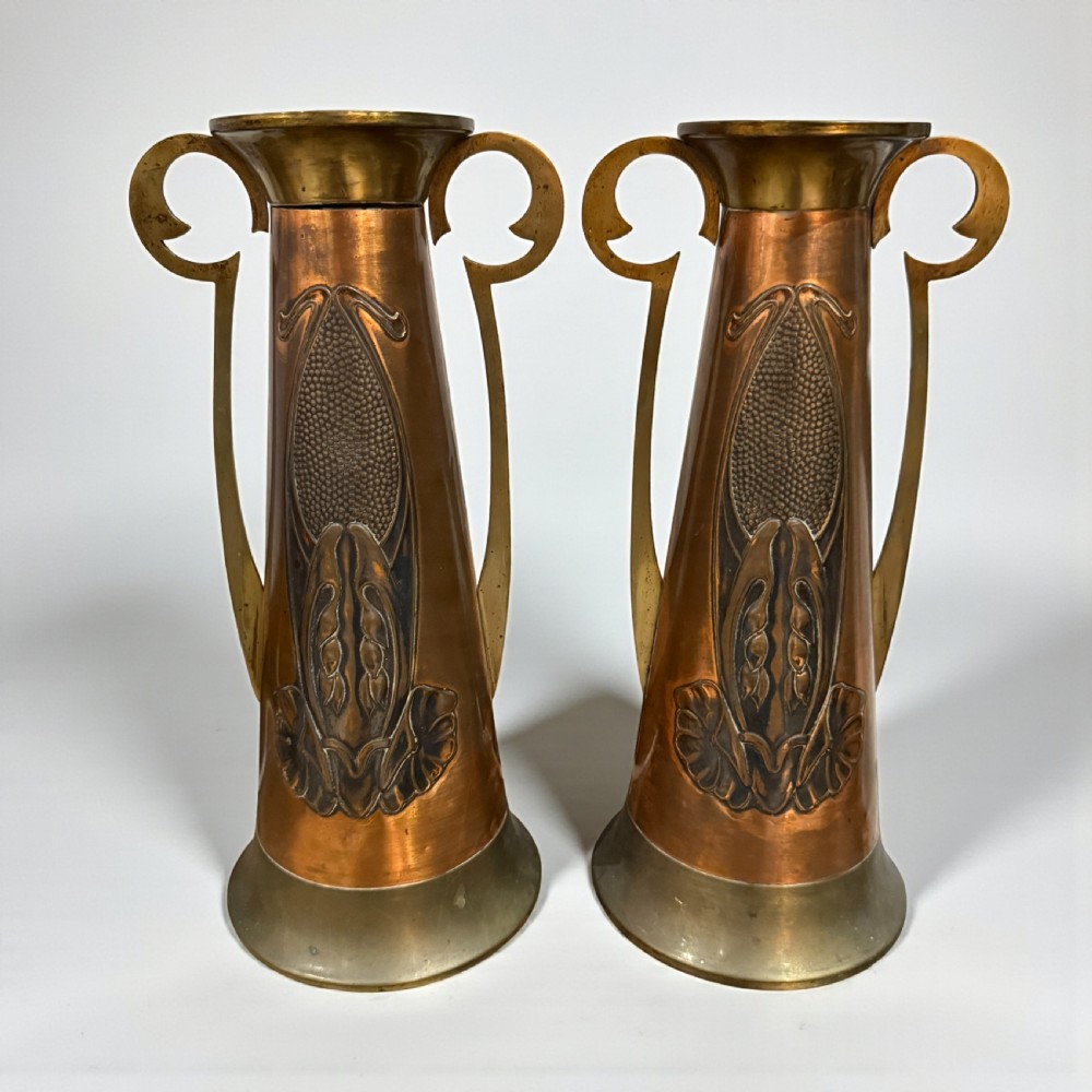 A pair of Arts and Crafts tapered copper vases featuring embossed seed pod motifs and sinuous brass handles.