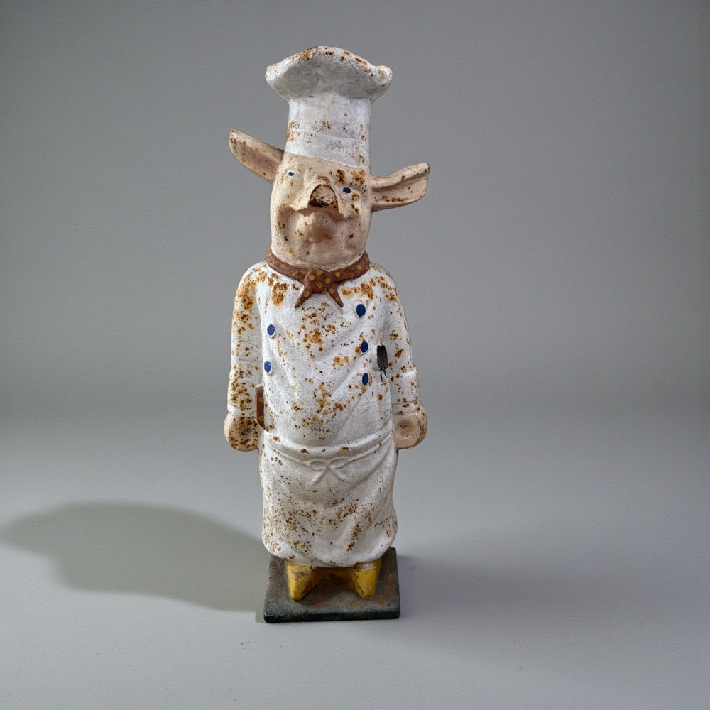 A standing painted cast iron pig figure wearing a white chef hat, apron, and neckerchief with surface rust patination.