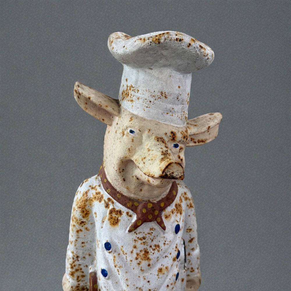 A standing painted cast iron pig figure wearing a white chef hat, apron, and neckerchief with surface rust patination. View II