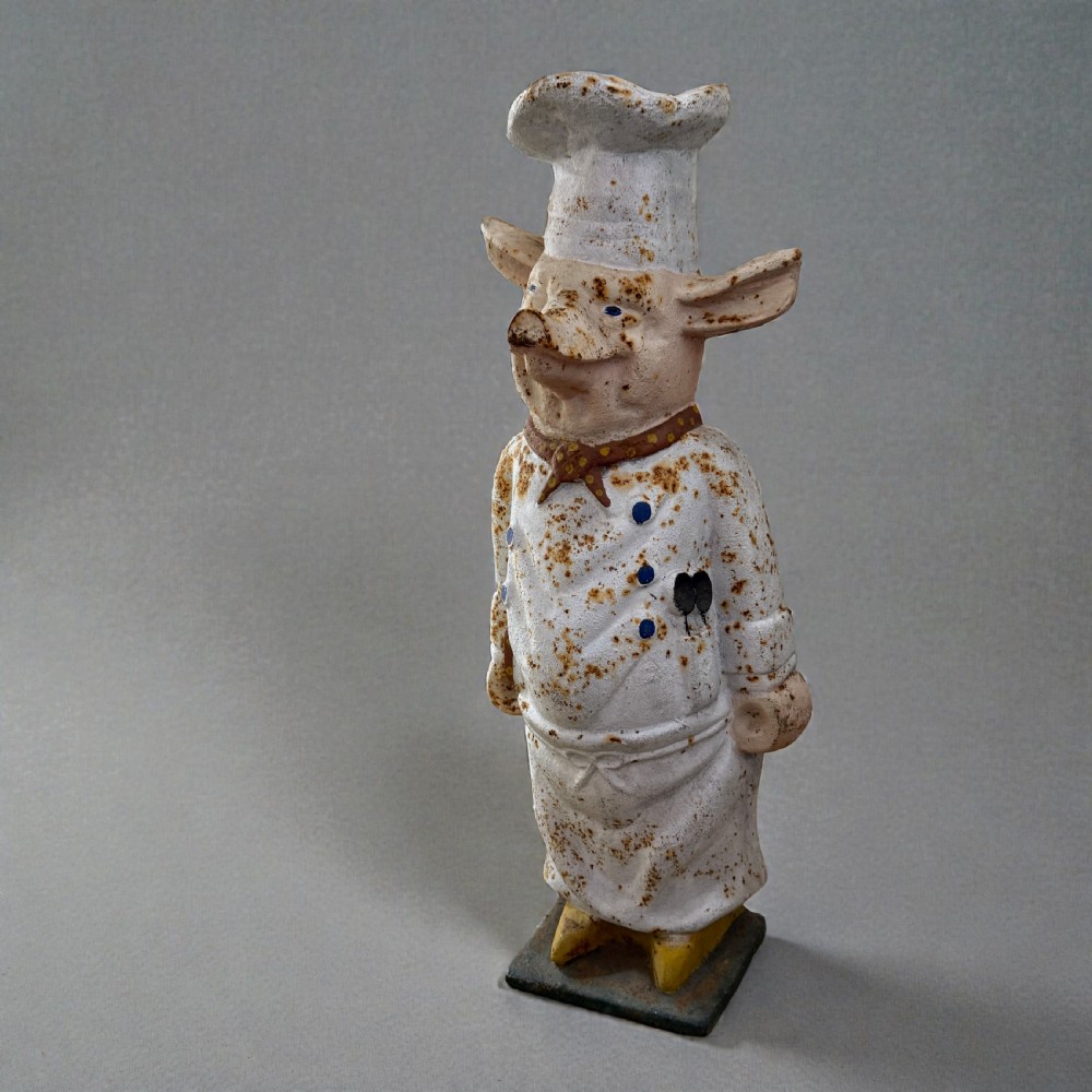 A standing painted cast iron pig figure wearing a white chef hat, apron, and neckerchief with surface rust patination. View III