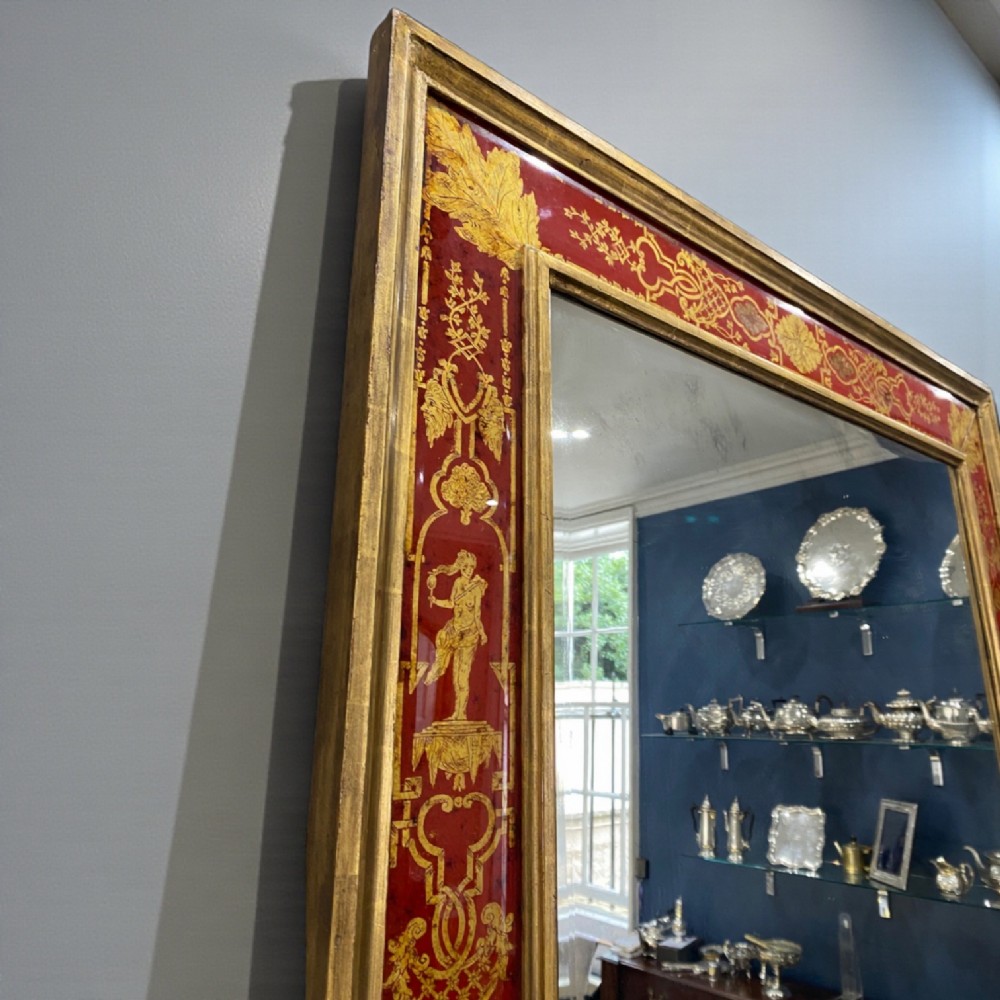 A rectangular wall mirror featuring a giltwood frame surrounding a broad red verre eglomise glass border decorated with gold scrollwork and classical figures. View II