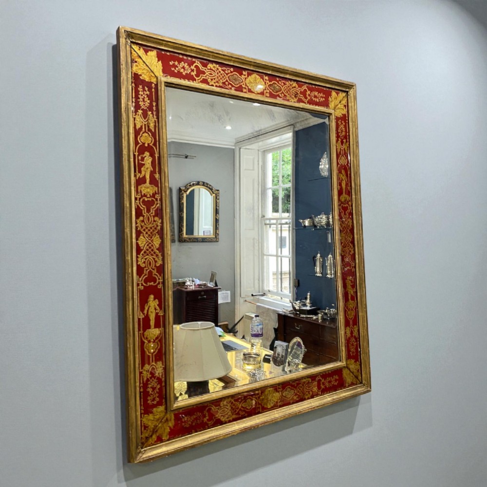 A rectangular wall mirror featuring a giltwood frame surrounding a broad red verre eglomise glass border decorated with gold scrollwork and classical figures.