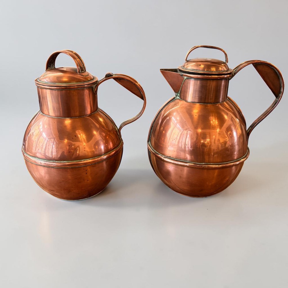 A pair of round copper Jersey milk jugs featuring fitted lids with top loop handles and wide pouring spouts.