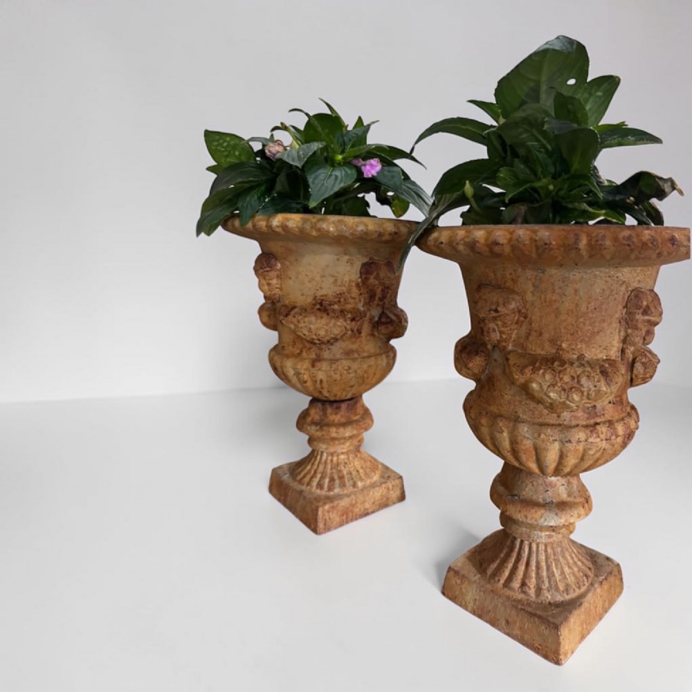 A pair of weathered brown cast iron garden urns featuring cherub reliefs on pedestal bases. View V