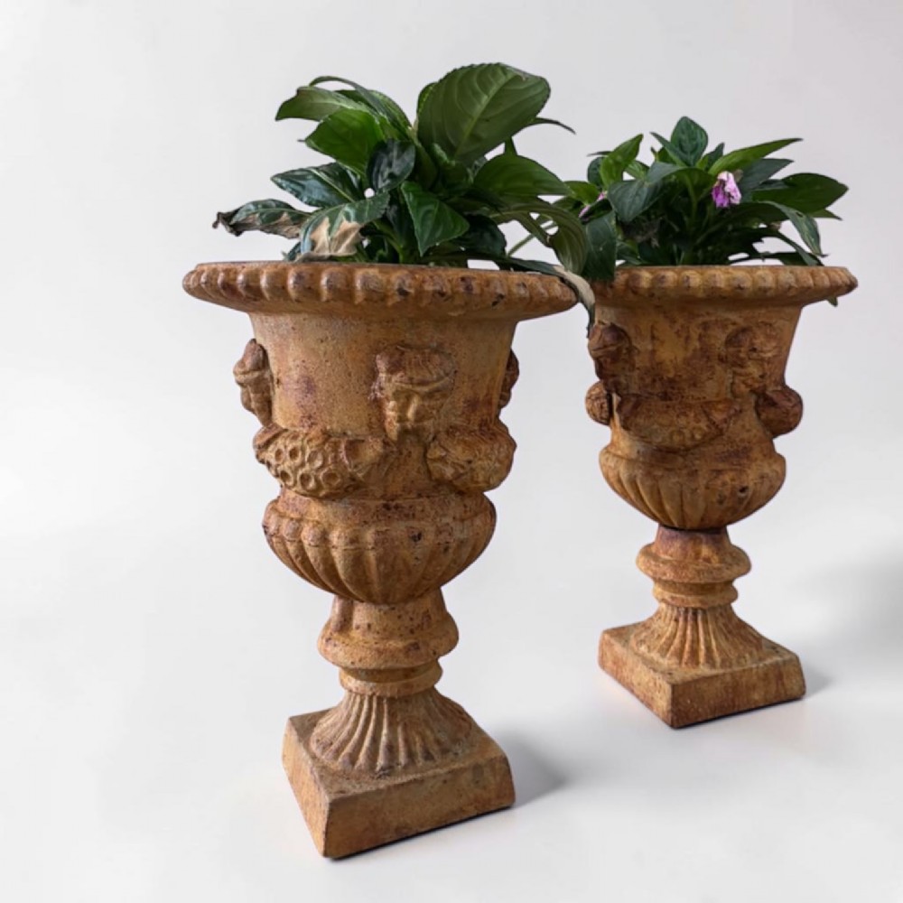 A pair of weathered brown cast iron garden urns featuring cherub reliefs on pedestal bases. View VI