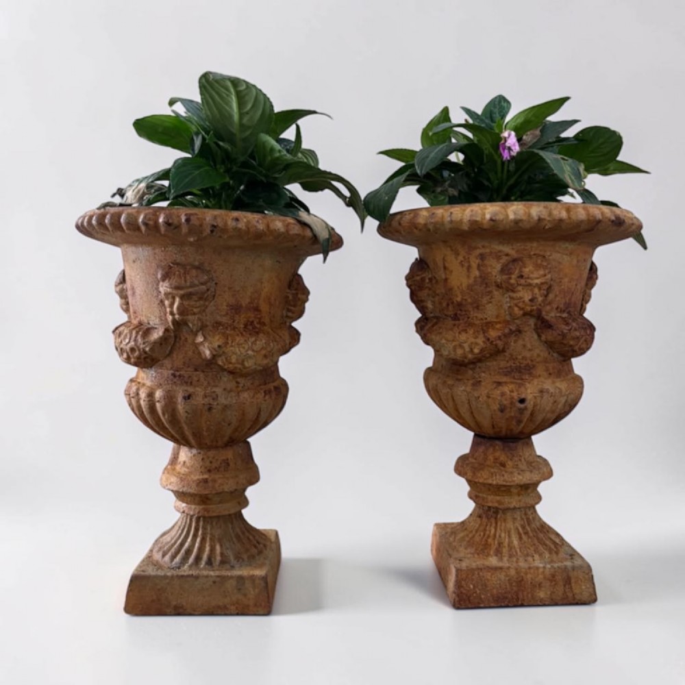 A pair of weathered brown cast iron garden urns featuring cherub reliefs on pedestal bases.