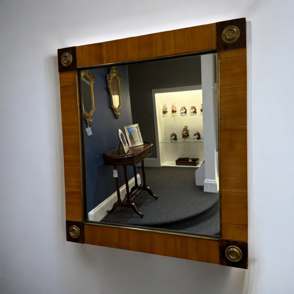 A square Biedermeier wall mirror featuring a warm brown wood veneer frame with dark corner blocks adorned with circular brass mounts.