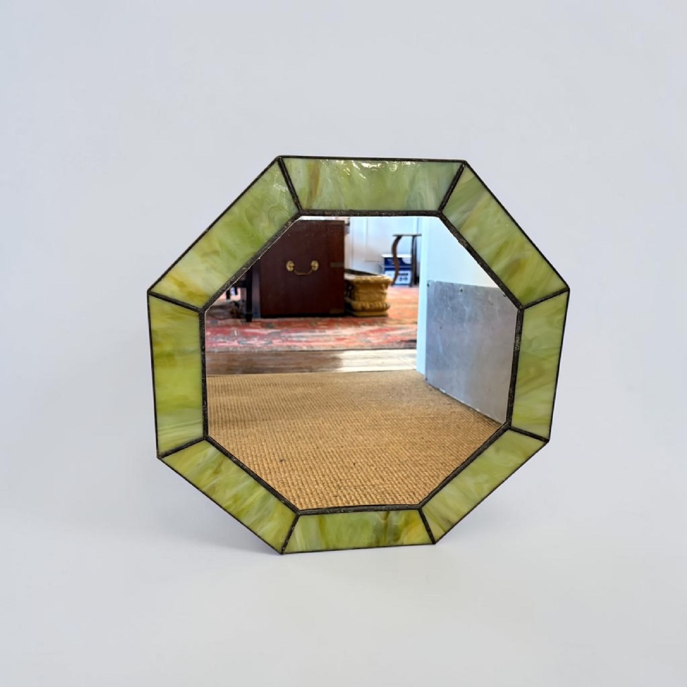 An octagonal wall mirror featuring a geometric border crafted from panels of variegated green stained glass joined with dark soldered seams.