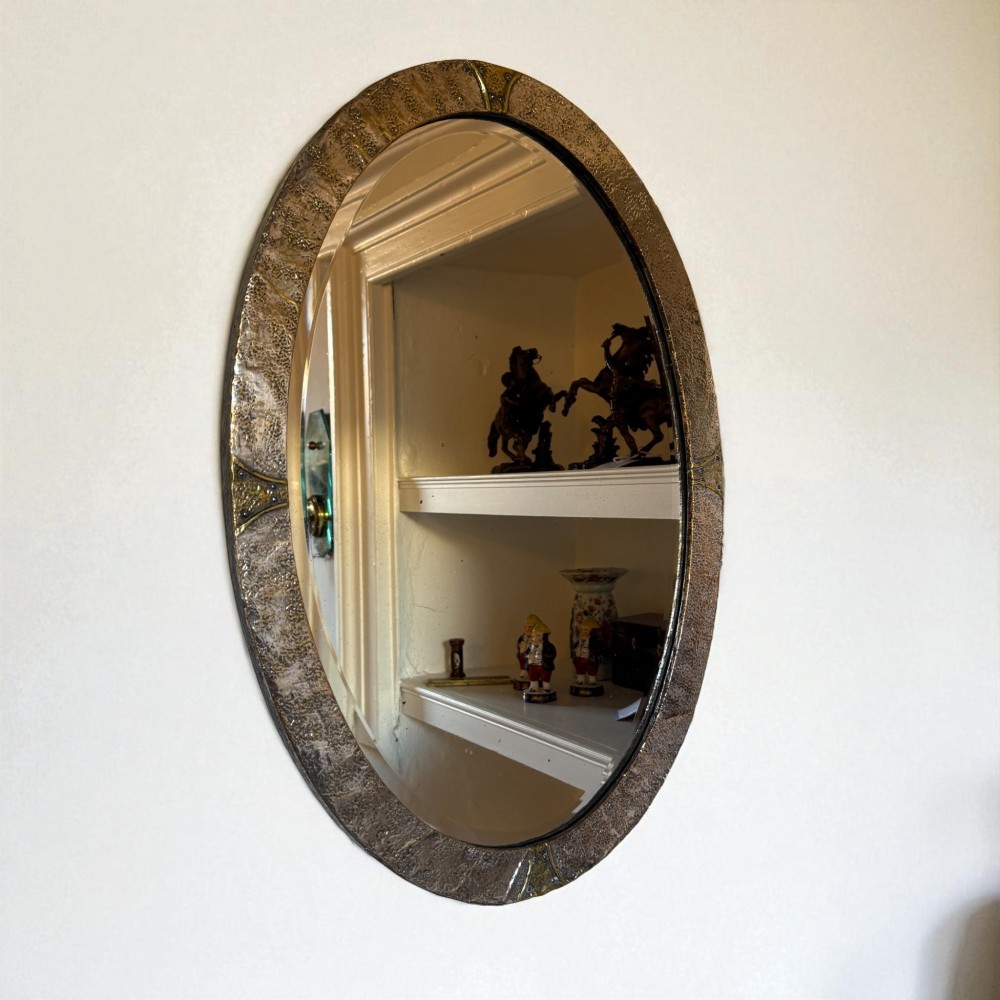 An oval Arts and Crafts wall mirror with a hand-hammered metal frame, decorative corner accents, and a bevelled mirror plate.
