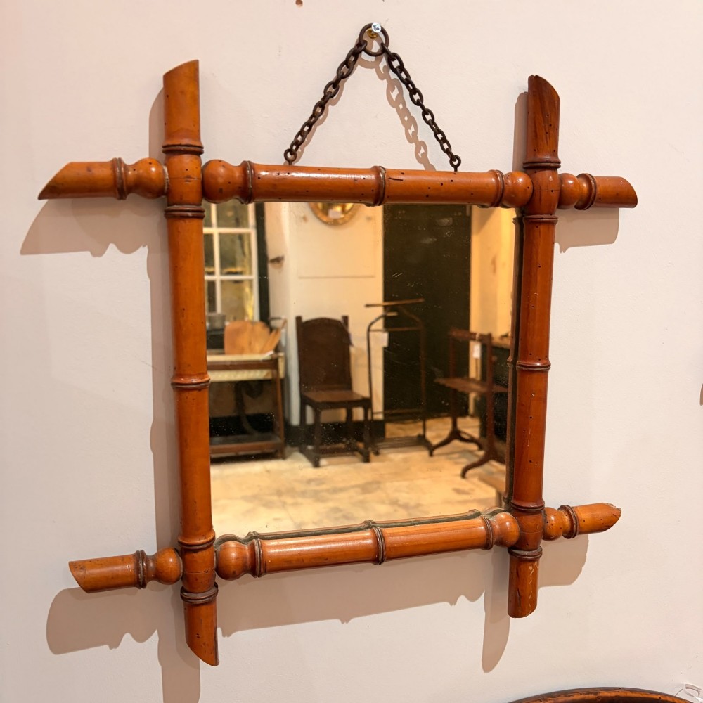 A square wall mirror framed in warm brown turned wood carved to resemble bamboo with extended corner joints and a metal suspension chain.