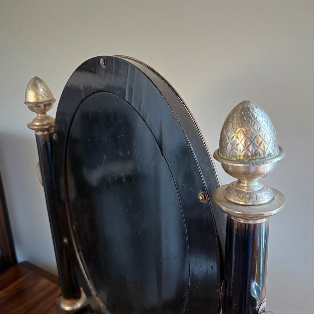 An antique 19th-century French ebonised toilet mirror featuring an oval glass frame suspended between twin columns with silvered finials and an arched base. View IV