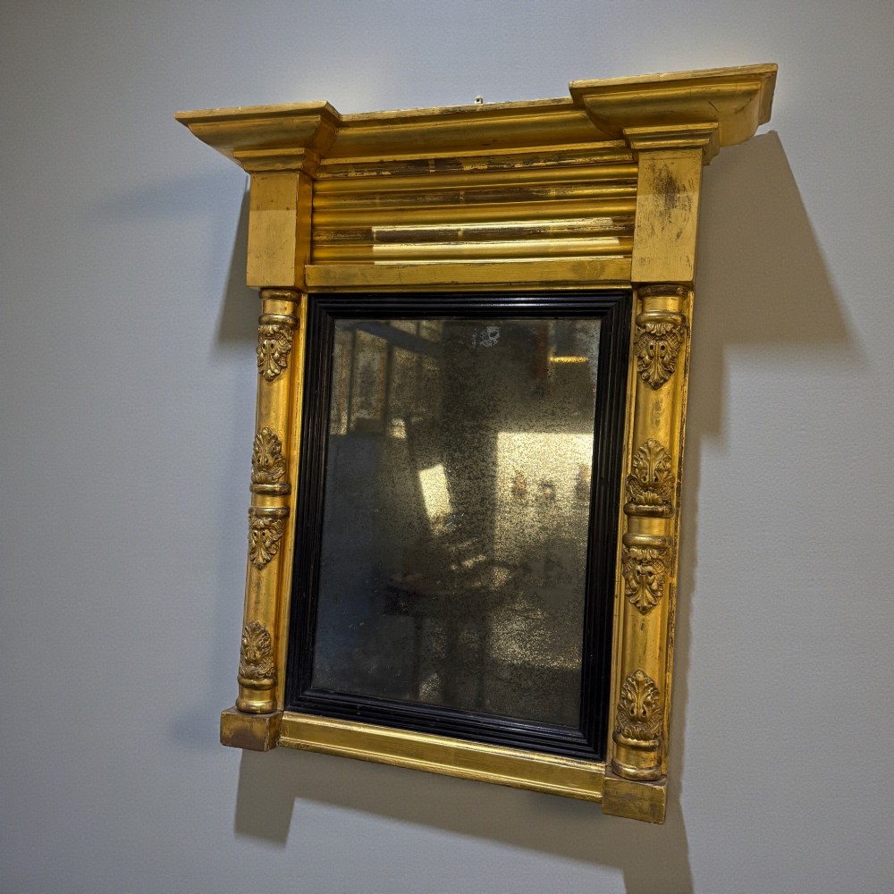A 19th-century giltwood wall mirror featuring an architectural breakfront cornice, acanthus leaf carved column pilasters, an ebonised inner slip, and foxed mirror glass.