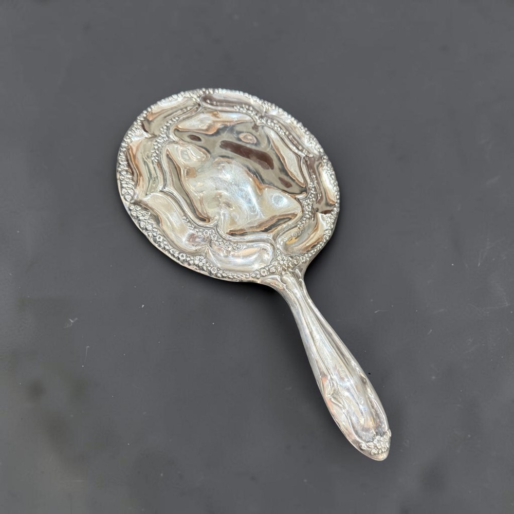 An antique silver hand mirror featuring embossed Art Nouveau scrollwork and a floral border around the oval metal casing.