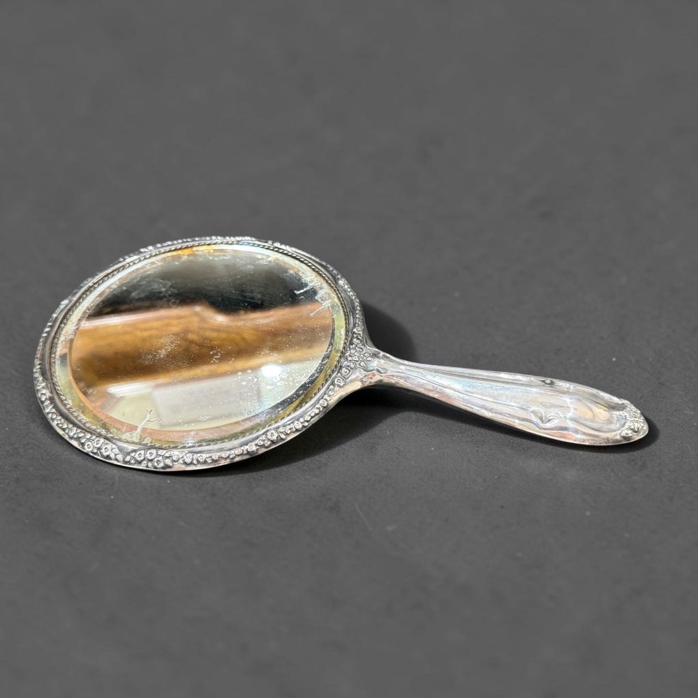 An antique silver hand mirror featuring embossed Art Nouveau scrollwork and a floral border around the oval metal casing. View II