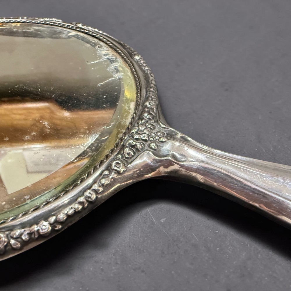 An antique silver hand mirror featuring embossed Art Nouveau scrollwork and a floral border around the oval metal casing. View III