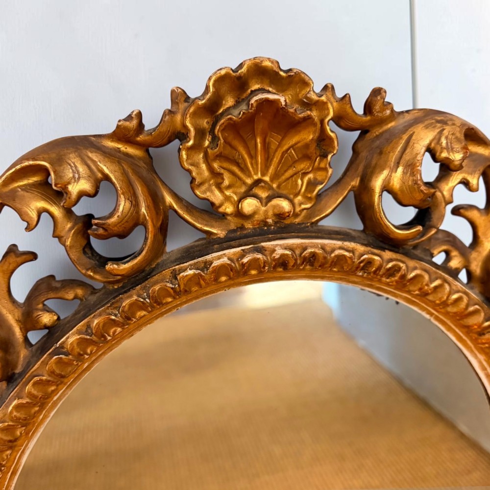 An oval mirror surrounded by an ornate gold gilt Florentine frame featuring carved acanthus leaf scrollwork and shell motifs. View II