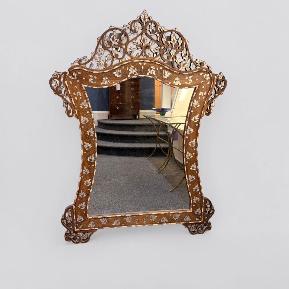 A Syrian wall mirror featuring a shaped dark wood frame with detailed mother-of-pearl floral inlays and an openwork pierced cresting.
