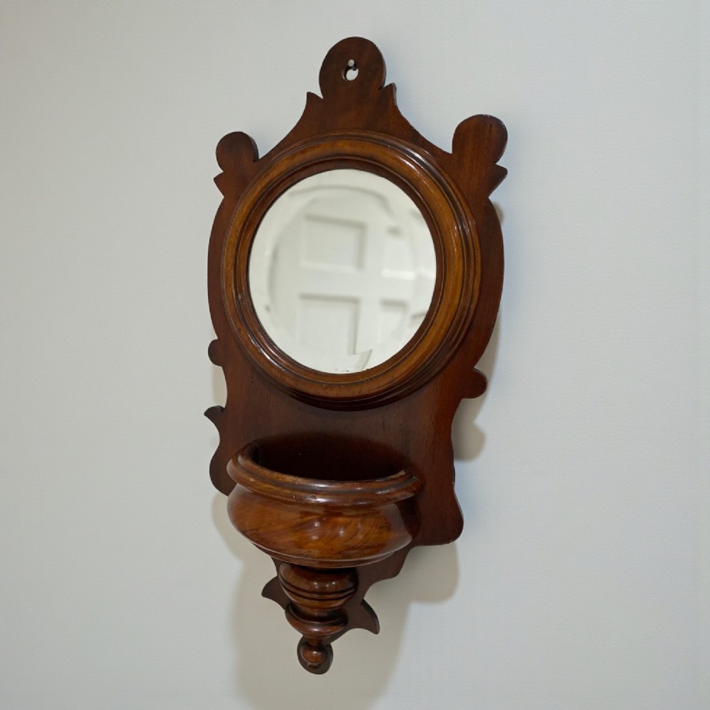 A wall-mounted Victorian mahogany bracket mirror featuring a circular mirror in a moulded frame above a carved wooden pocket and turned bottom finial. View II