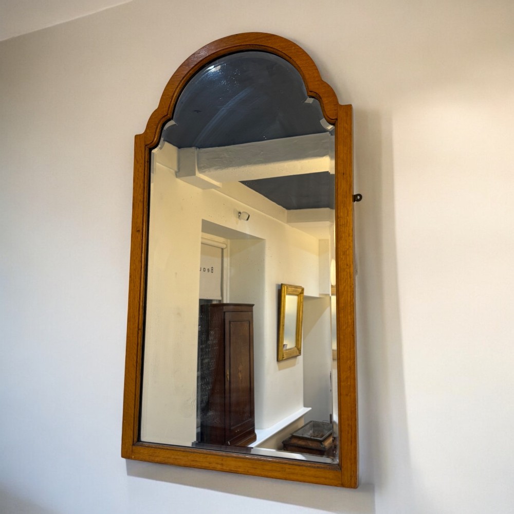 An upright rectangular wall mirror with a warm oak wooden frame and a shaped arched crest at the top.