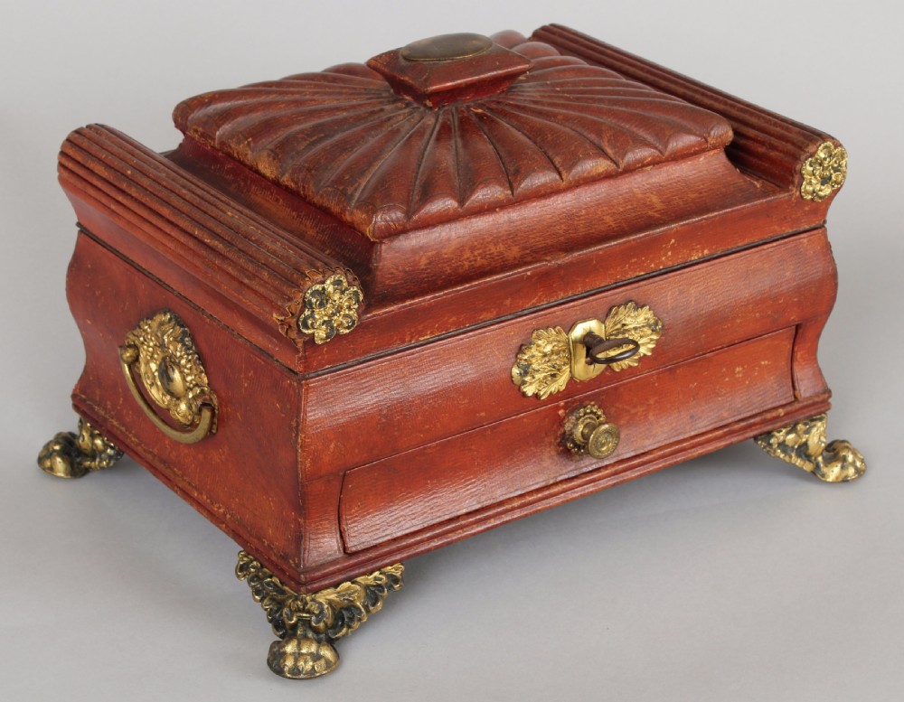 A Regency period sarcophagus-form work-box bound in red Russia leather with gilt-brass paw feet, side handles, and escutcheon.