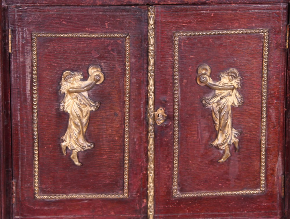An early nineteenth-century table-cabinet bound in deep red tooled leather, featuring fluted column corner posts, classical gilt-brass figure mounts on the twin doors, and dolphin side handles. View VI
