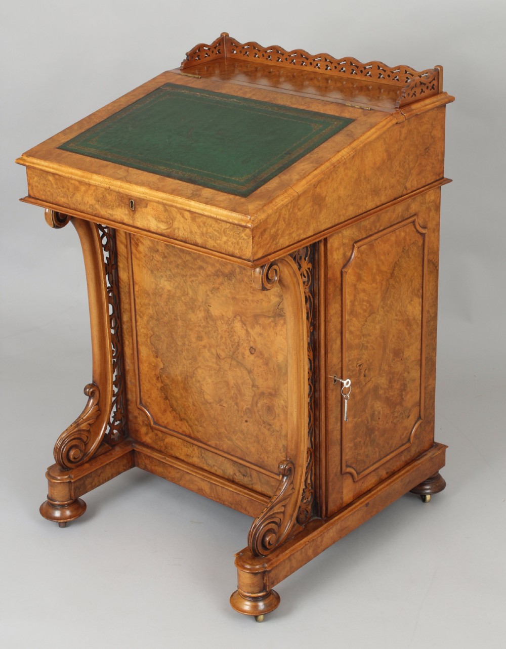 A Victorian burr walnut Davenport desk featuring a sloped green leather writing top, carved fretwork gallery, and side cabinet door on brass castors.