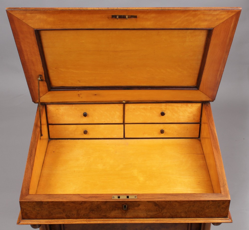 A Victorian burr walnut Davenport desk featuring a sloped green leather writing top, carved fretwork gallery, and side cabinet door on brass castors. View V