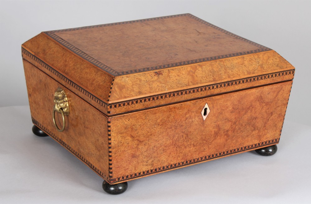 A Regency period sarcophagus-shaped work box crafted from golden birdseye maple with geometric ebony inlay, brass lion mask ring handles, and ball feet.