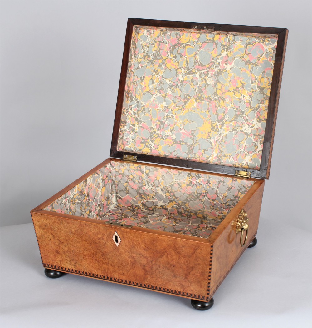 A Regency period sarcophagus-shaped work box crafted from golden birdseye maple with geometric ebony inlay, brass lion mask ring handles, and ball feet. View III