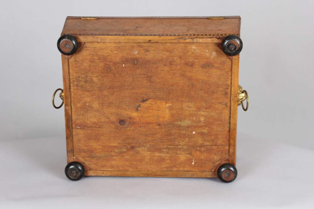 A Regency period sarcophagus-shaped work box crafted from golden birdseye maple with geometric ebony inlay, brass lion mask ring handles, and ball feet. View V