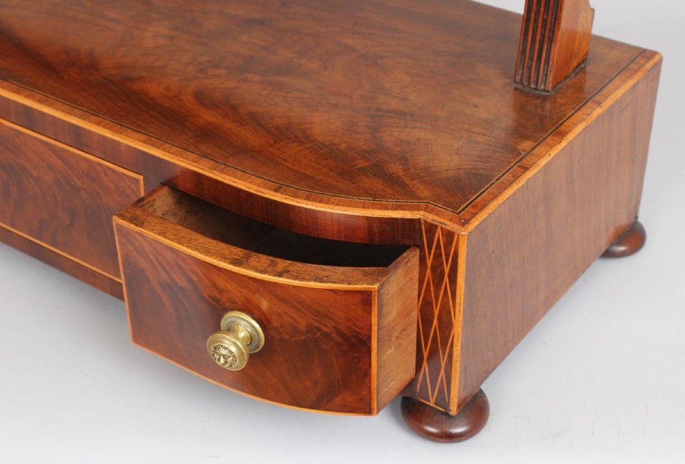 A George IV mahogany toilet mirror featuring an adjustable rectangular glass plate supported by fluted uprights over a bow-fronted drawer base. View II