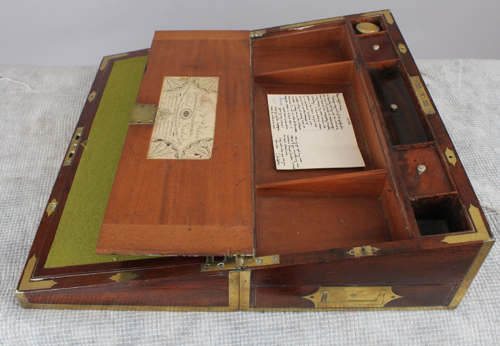 An open antique mahogany writing box with brass corner mounts, revealing a green baize writing surface, internal pen tray compartments, and a stationers paper label inside the lid.