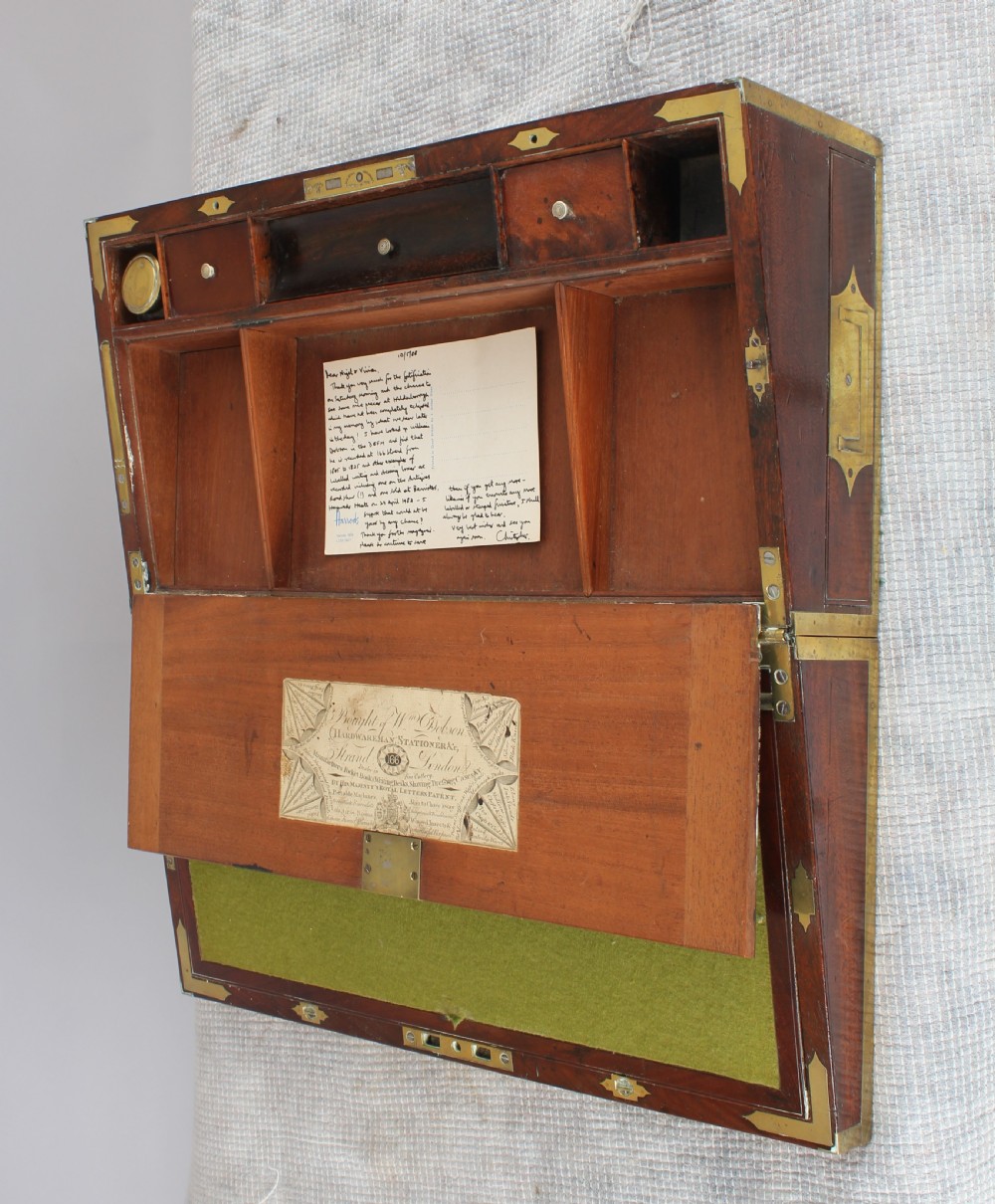 An open antique mahogany writing box with brass corner mounts, revealing a green baize writing surface, internal pen tray compartments, and a stationers paper label inside the lid. View IV