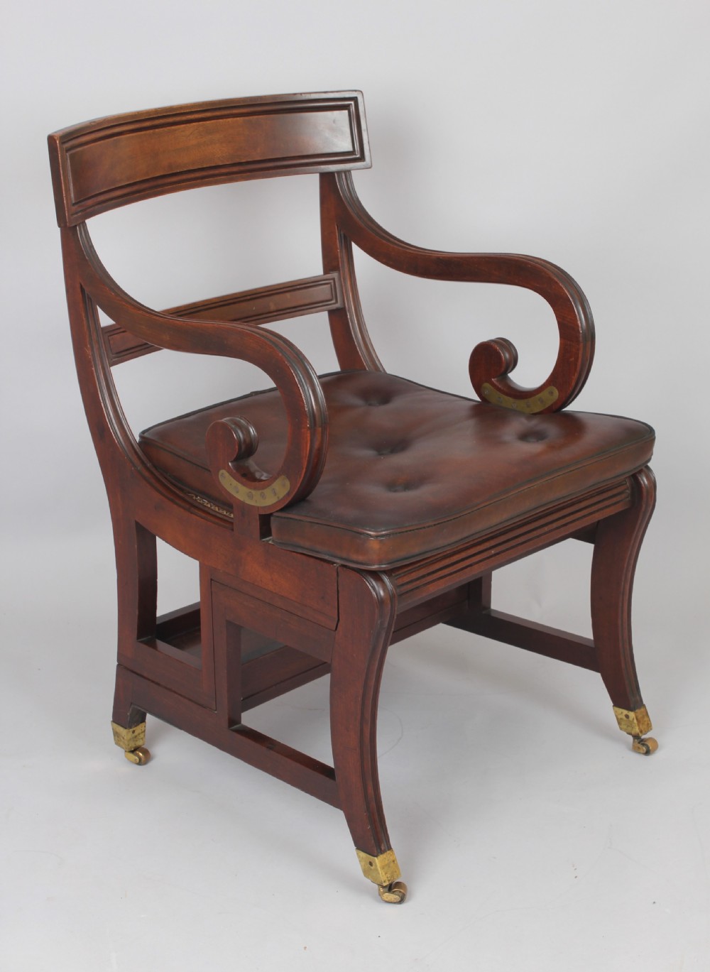 A Regency period mahogany metamorphic library chair with scroll armrests, a buttoned leather seat, and brass castors.