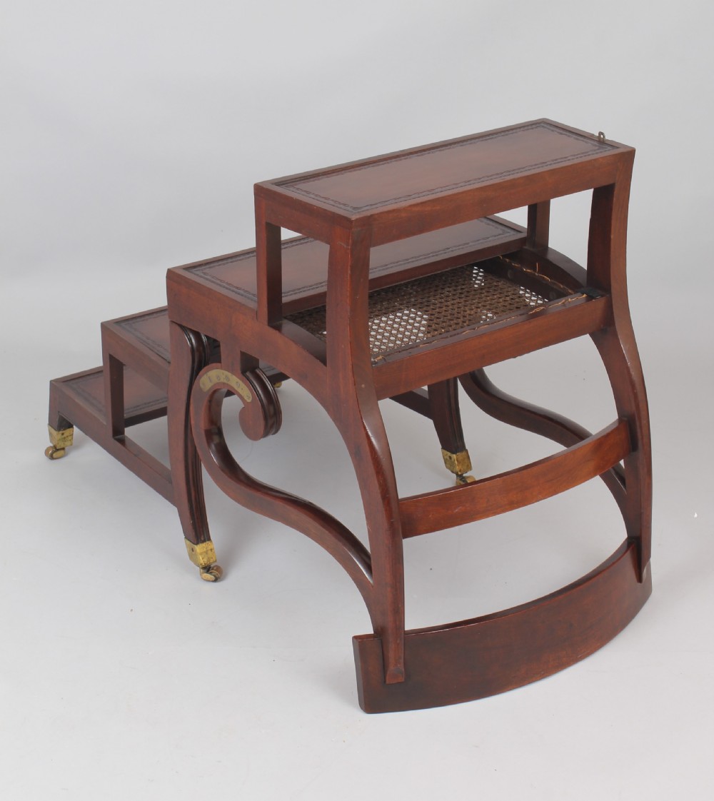 A Regency period mahogany metamorphic library chair with scroll armrests, a buttoned leather seat, and brass castors. View II
