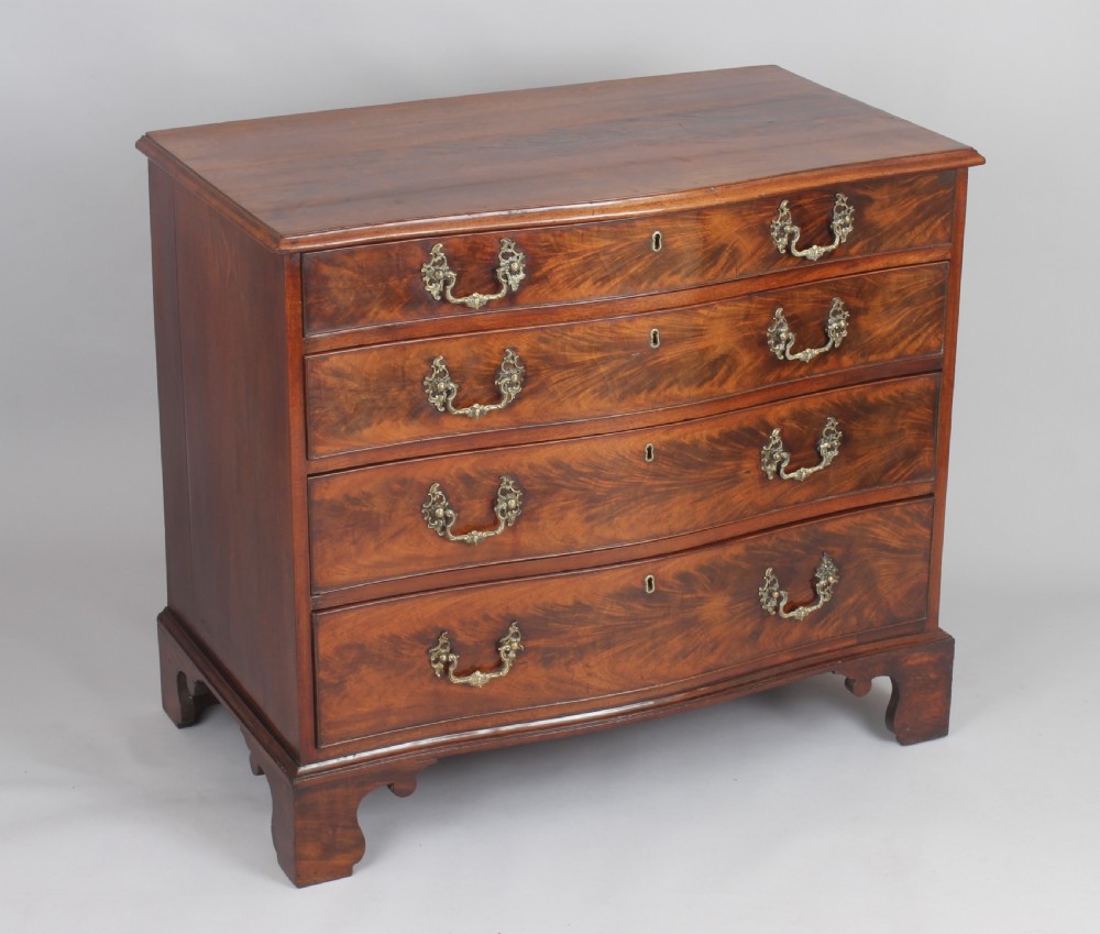A Georgian style warm brown mahogany serpentine chest of drawers featuring four graduated drawers with rococo brass handles and bracket feet.