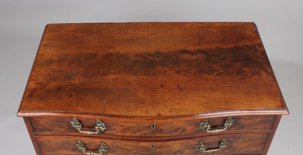 A Georgian style warm brown mahogany serpentine chest of drawers featuring four graduated drawers with rococo brass handles and bracket feet. View V